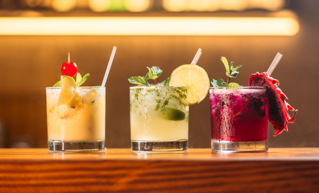 Three colorful cocktails in glasses garnished with fruit and mint, displayed on a bar counter