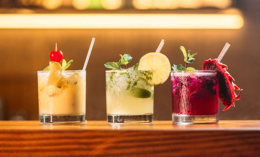 Three colorful cocktails in glasses garnished with fruit and mint, displayed on a bar counter